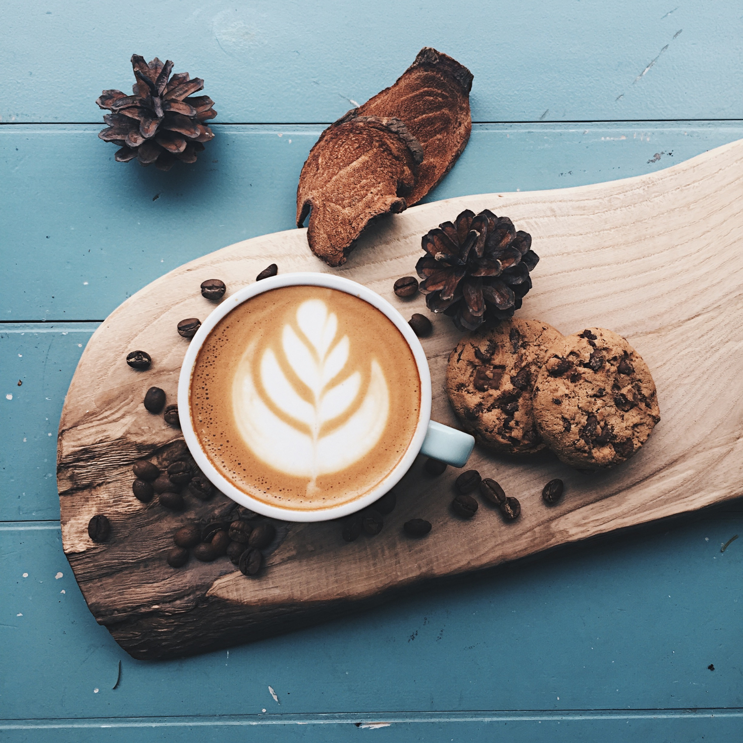 A cup of latte with intricate latte art on a wooden tray, accompanied by chocolate chip cookies, coffee beans, and pinecones, creating a cozy and inviting coffee experience.